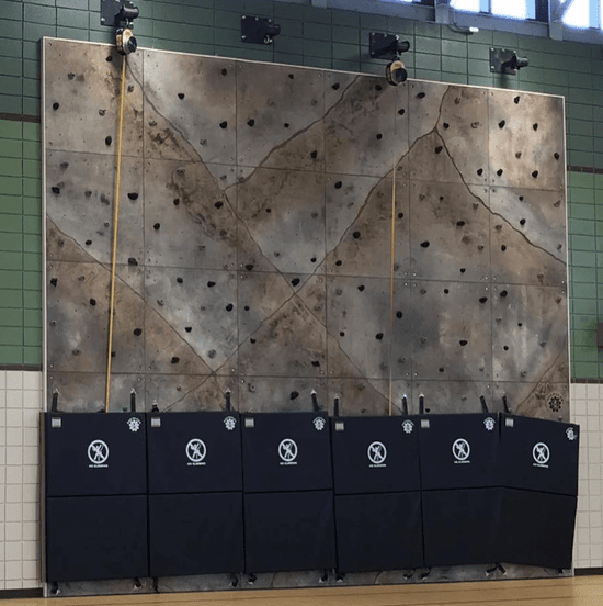 A moderately sized custom sculpted gym rock (concrete) flat rock climbing surface against a wall, with four belay stations, two auto belays, and folding security mats lined up at the bottom.