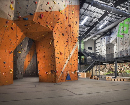 Indoor rock climbing gym with an orange and gray tone rock climbing lead cave with top rope and auto belay options to the right side, and a neatly laid carpet bonded foam floor.