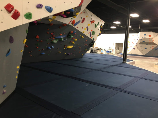 Rock climbing gym bouldering cave to the left, walking path to the right, and a neatly laid out black fabric covered bouldering foam flooring in the middle and under the boulder cave.