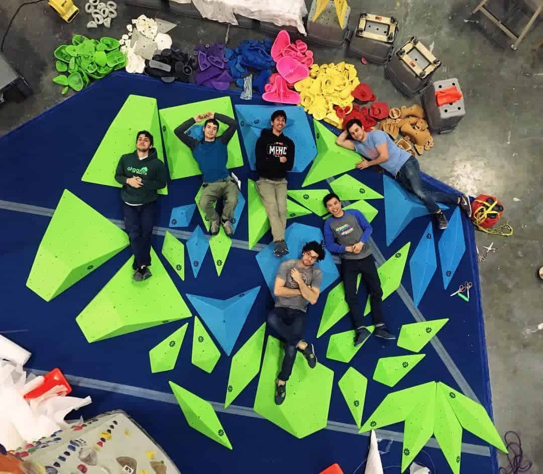 People laying down on rock climbing volumes spread out on the padded flooring of a climbing gym with climbing holds in piles around the flooring perimeter.
